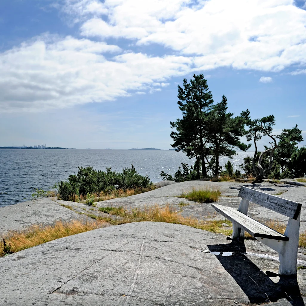 A wooden bench on a rocky shore overlooking the calm blue sea under a cloudy sky.