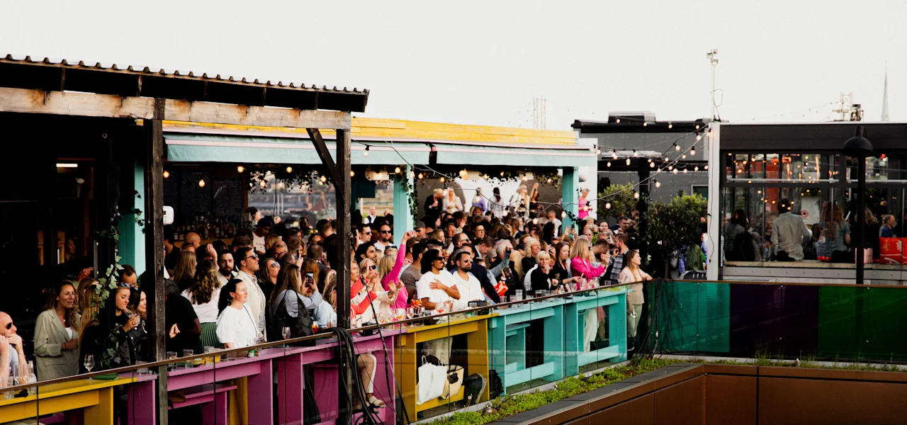 En livlig folkmassa som njuter av en social sammankomst på en färgglad takterrass med ljusslingor.