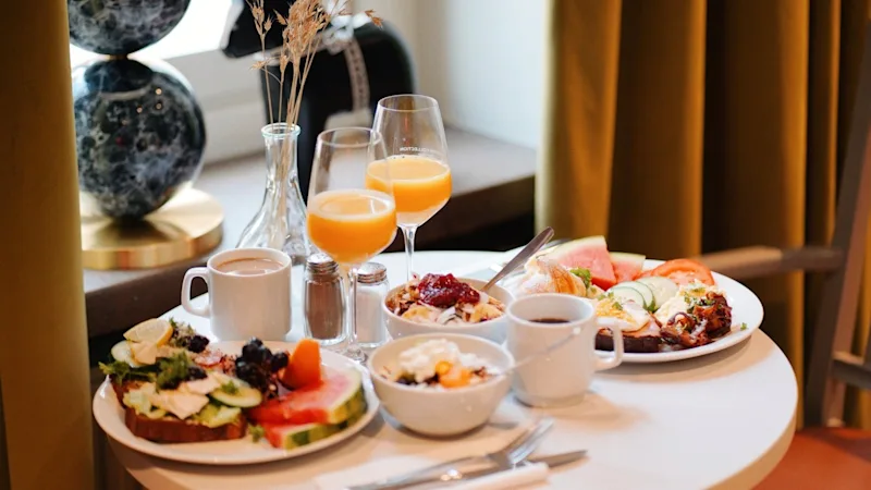 Ett frukostbord med tallrikar med blandade frukter och bakverk, glas med apelsinjuice och kaffekoppar i en mysig, mjukt upplyst matsal nära fönster med gardiner.