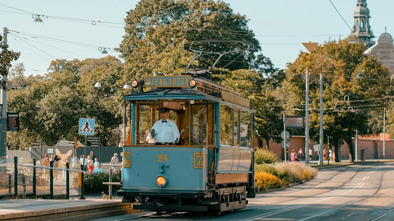 A light blue tram with a driver in uniform travels down a street on a sunny day. DJURGARDEN is visible on the tram.