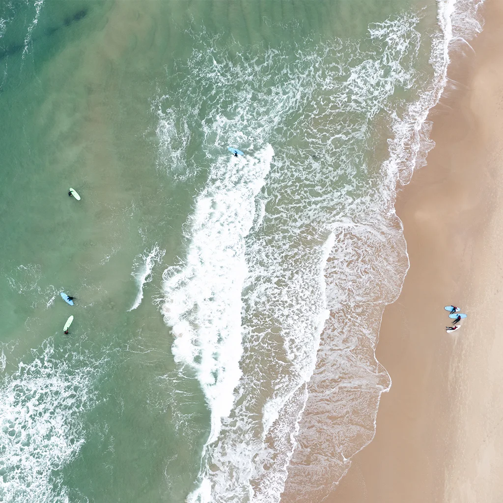 Ilmakuva hiekkarannasta turkoosine merivesineen ja murtuvine aaltoineen. Useat ihmiset surffaavat ja nauttivat vedestä.