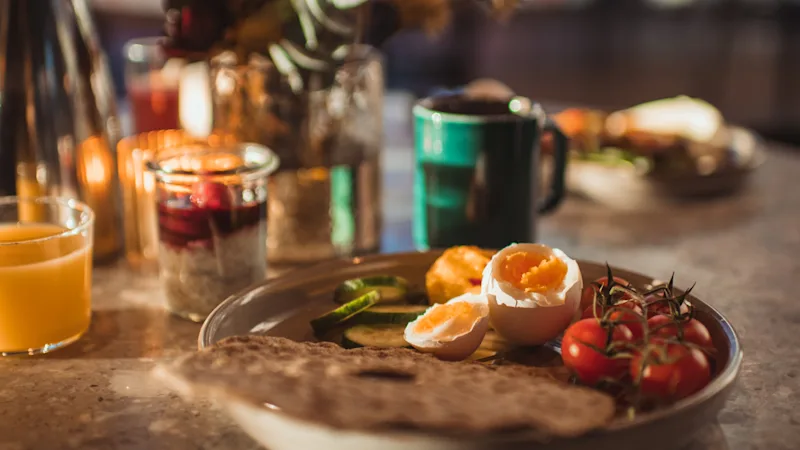 Frukosttallrik med löskokta ägg, tomater och gurkskivor står på ett bord. Solen strömmar in genom ett fönster och belyser en varm, mysig matsal med en mugg och juice.