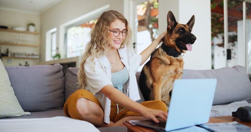 A smiling woman sits cross-legged on the couch with her laptop, one hand resting on her adult German shepherd beside her.