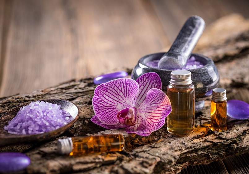 Aromatherapy setup with essential oil bottles, purple bath salts, and a pink orchid flower on rustic wood, prompting questions like are essential oils safe for cats and which essential oils are safe for cats, while emphasizing the importance of using pet safe essential oils and cat safe essential oils for diffuser use.