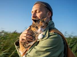 A man in a gray beanie and olive green jacket embraces his calico cat outdoors, eyes closed in a moment of pure contentment as tall grasses sway against a brilliant blue sky behind them. This outdoor excursion captures the growing trend of people wondering can cats be service animals as they bring their feline companions into more aspects of daily life.