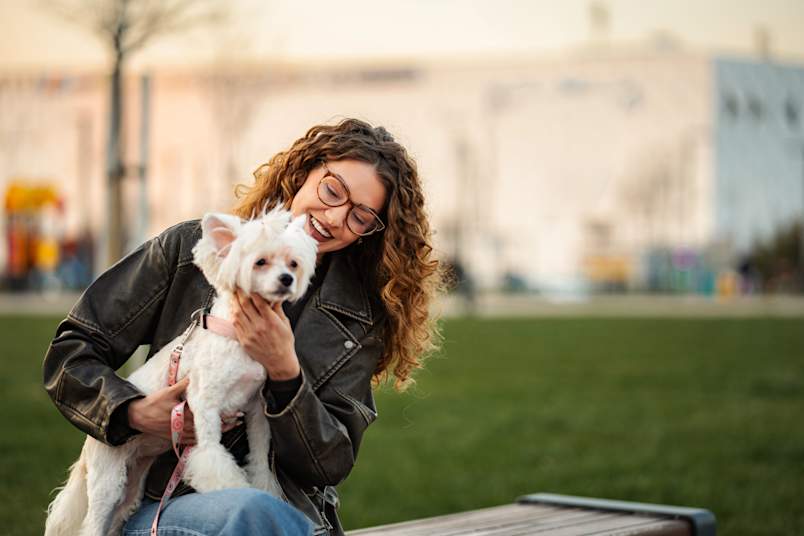 A young woman in a leather jacket sits on a park bench and laughs while cradling her small white dog, the kind of bond that makes choosing the perfect name feel important.
