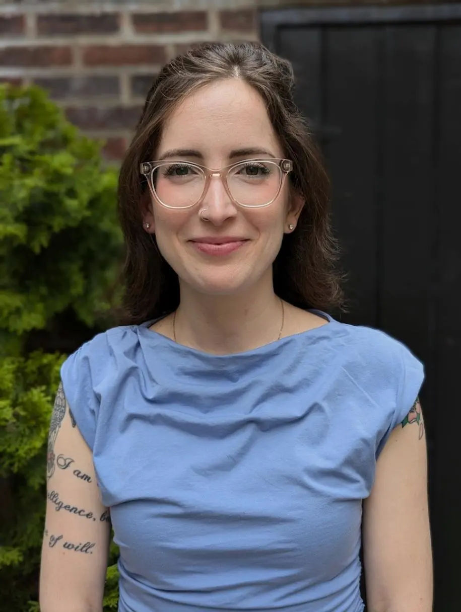 Therapist Gianna Damis LPC Portrait Therapist Gianna Damis wearing glasses and a blue top against a dark background, staring into the camera and smiling softly.