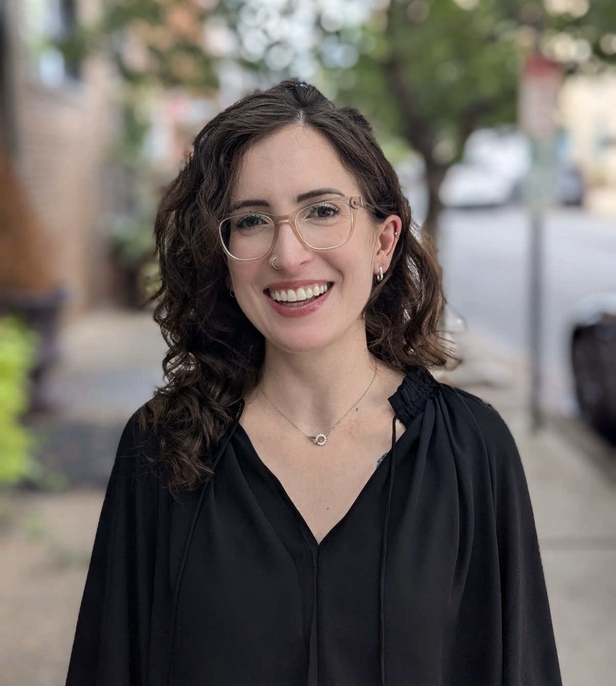 Therapist Gianna Damis LPC Portrait Therapist at Created Self Therapy, Gianna Damis, smiles warmly for a headshot, wearing glasses and a black top against the blurred background of a tree-lined street.