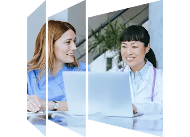 Two women in medical scrubs seated at a table, working together on a laptop