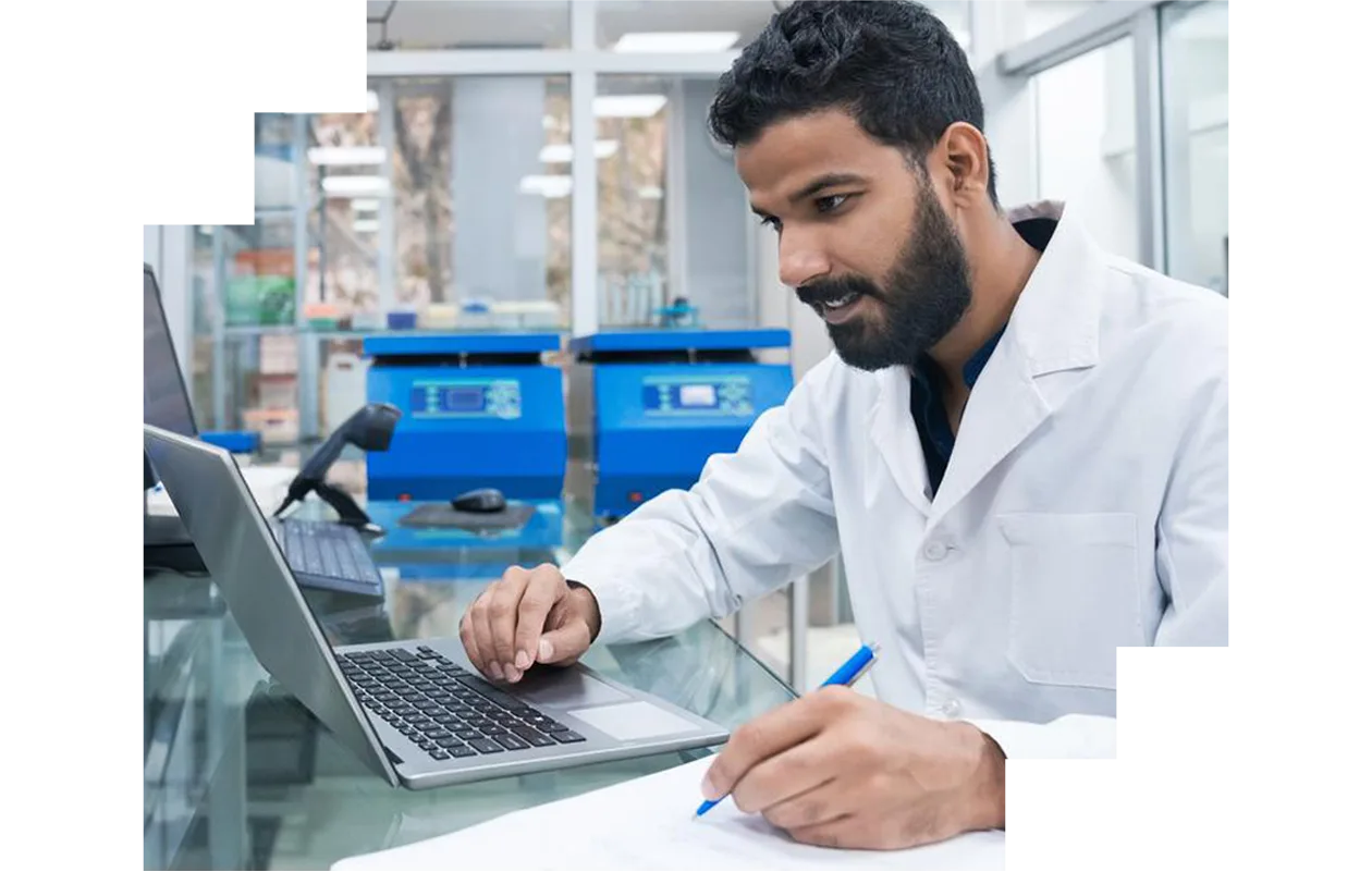 Young male biochemist using laptop while writing on clipboard at laboratory