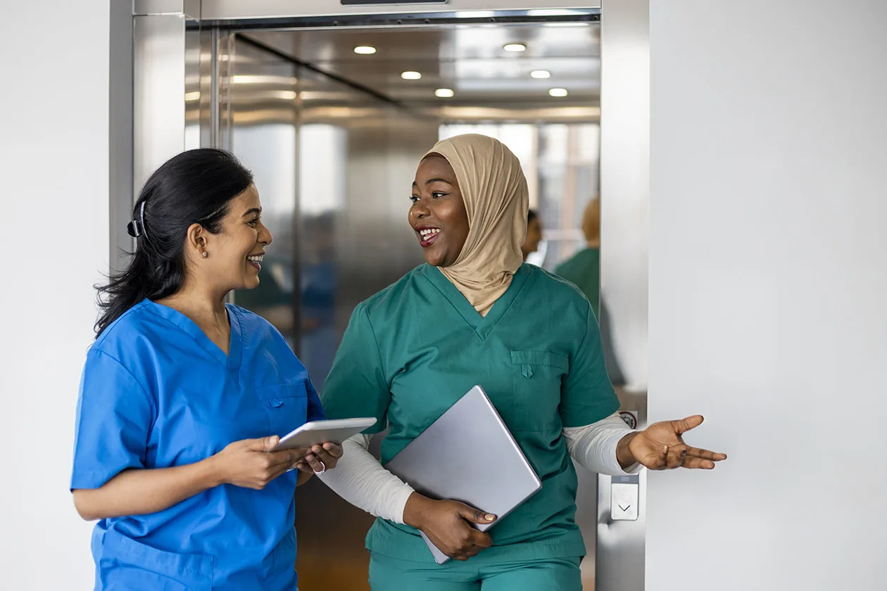 Two female nurses smiling and talking while walking by an elevator.