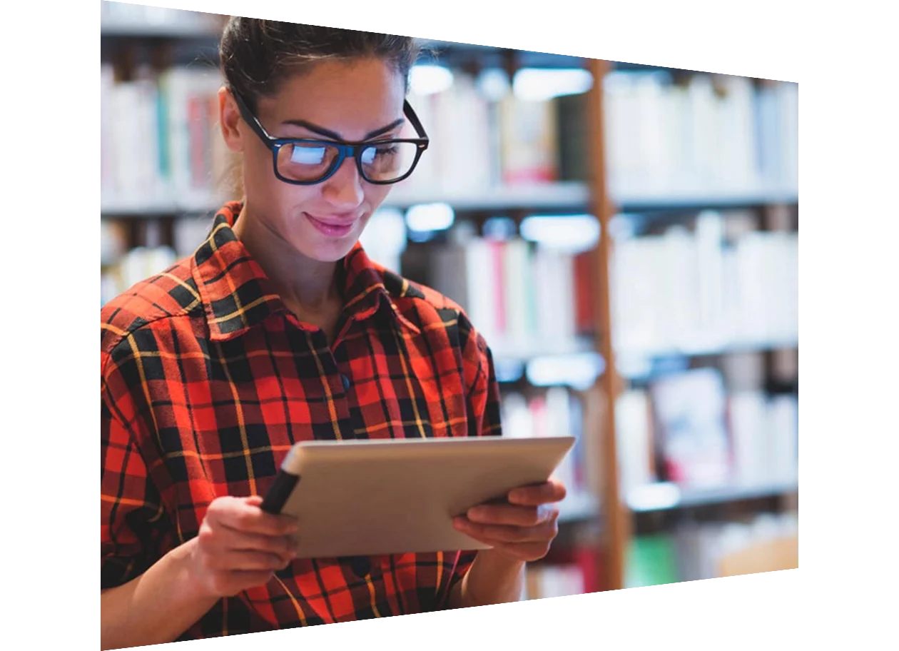 A young woman in a library wearing glasses looking down at a tablet