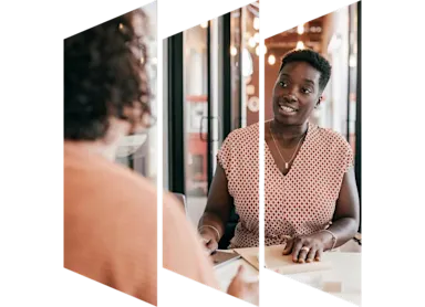 Two businesswoman sitting at desk and talking to one another.