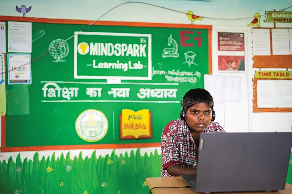 A young Indian boy wears headphones while looking at a laptop screen. He sits inside a classroom.
