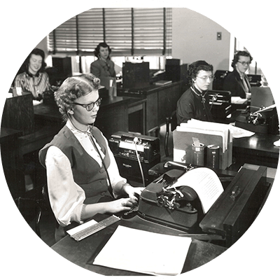 A black and white photo of P&G’s first blind employee, Jane Dotson, who was hired in January 1950 and worked in the transcribing department at the General Offices. She sits at a desk, typing on a typewriter amongst other women transcribers.