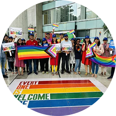 A large group of Indian men and women stand together in front of large Pride flag painted on the ground. They also are holding up numerous Pride flags.