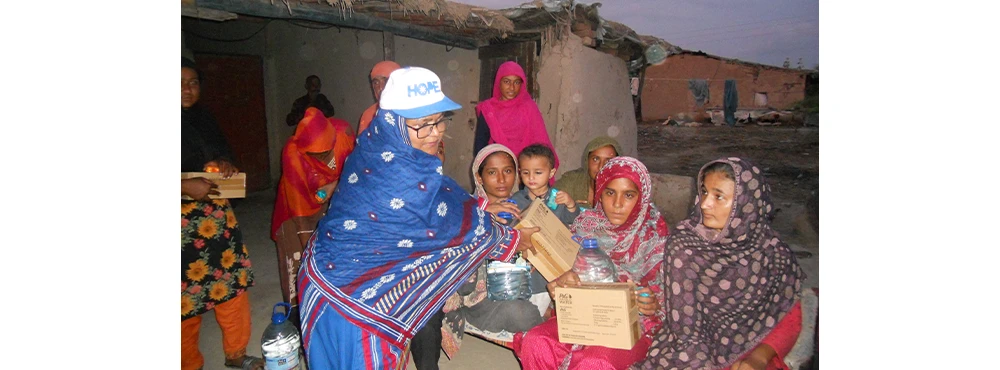 Women in scarves and traditional garments, including one in a white baseball hat, hold and exchange boxes of aid.