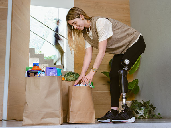 A woman with a prosthetic leg picks up bags of groceries outside her house.