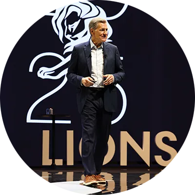 A man with short grey hair wearing a white collared shirt and a black suit. He stands on a stage with a black background.