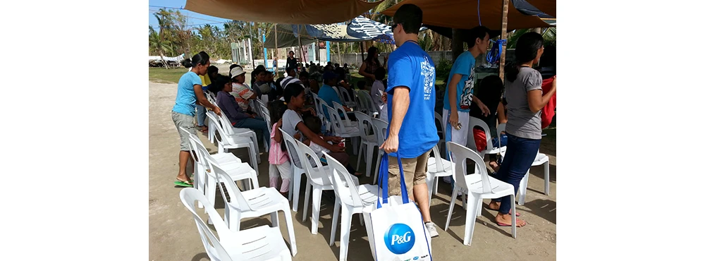 A man in a blue shirt holds a P&G branded bag while walking toward a seated group of community members awaiting support after a disaster.