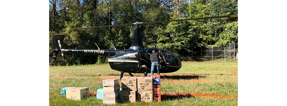 A black helicopter in an open field with a man nearby and several P&G disaster relief kits and products on the ground, ready to be loaded into the helicopter.