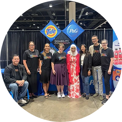 A group of people smiles in front of a conference booth display featuring P&G and Tide logos and multicolored text that says, “Disability Confident.”