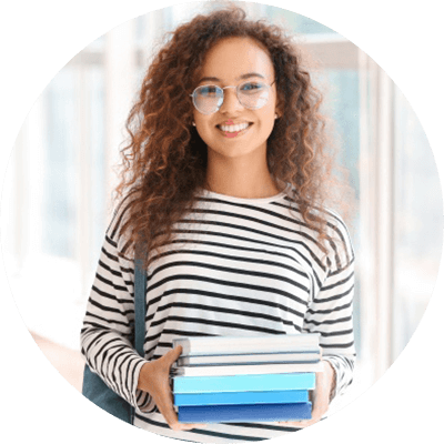 A young woman with long, brown curly hair and glasses smiles directly at the camera. She is wearing a black and white striped shirt and is holding a set of books.
