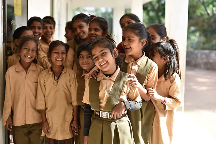 A group of 14 young Indian girls, dressed in brown and olive-green school uniforms, smile while gathered outside a classroom.
