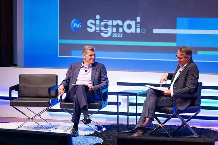 Two mean dressed in business suits sit on a stage for a conversation. A digital image in the background includes the blue and white Procter and Gamble logo and the Signal 2023 typeface logo.