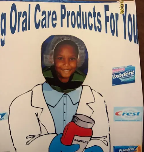 A small child sticking his head through a poster which says, “Oral Care Products for You”