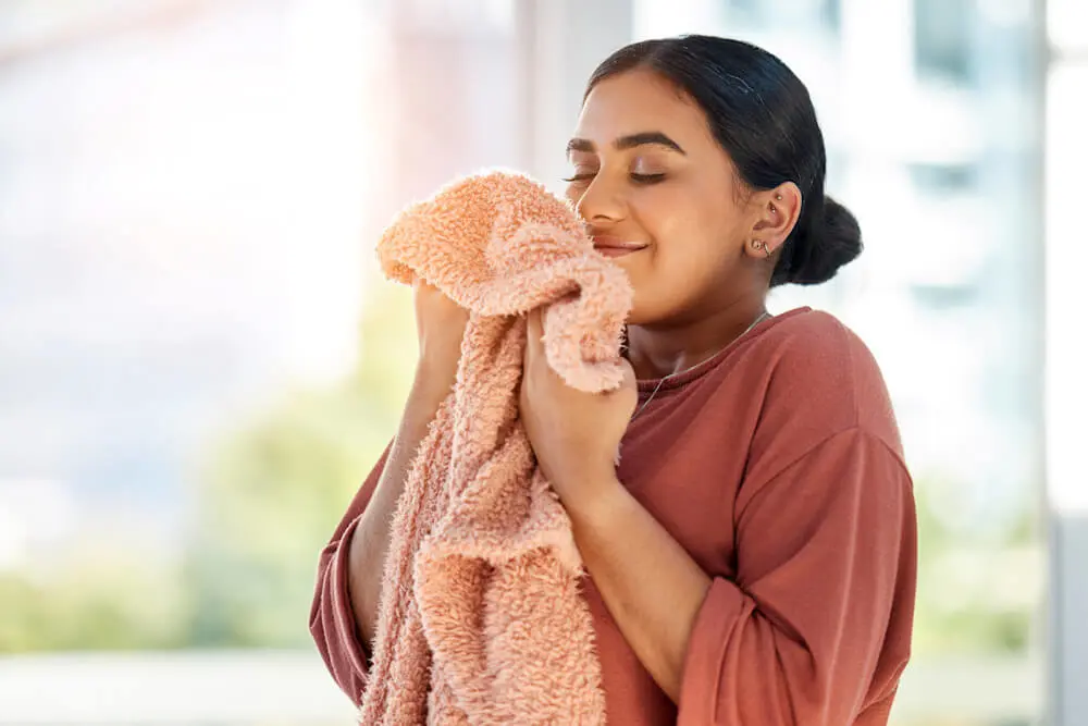 A woman with dark in a pink shirt holds up a pink colored blanket. She holds it up to face as she smells the fabric and smiles.