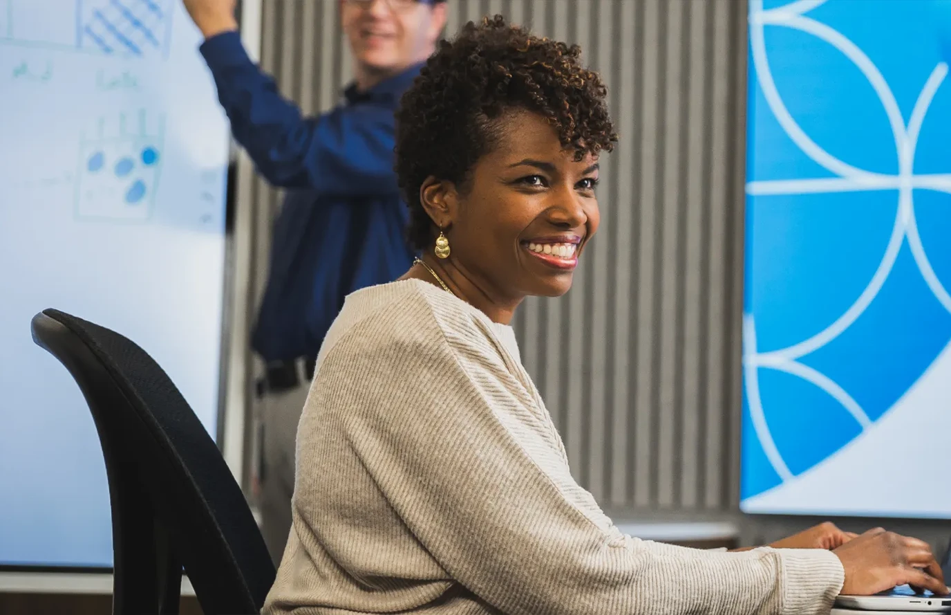 Woman seated at a computer while a man presents at a board in the background.