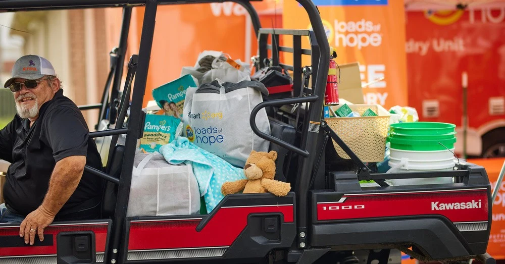 A man wearing sunglasses and a grey hat sits in a utility vehicle loaded with diapers, bags, buckets, and other relief supplies in front of an orange mobile disaster relief laundry unit.