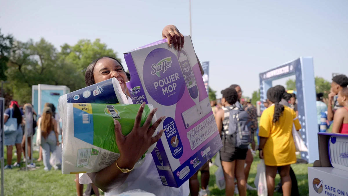 A black female student holds P&G brand products in her arms and smiles. She is in an outdoor college campus event.