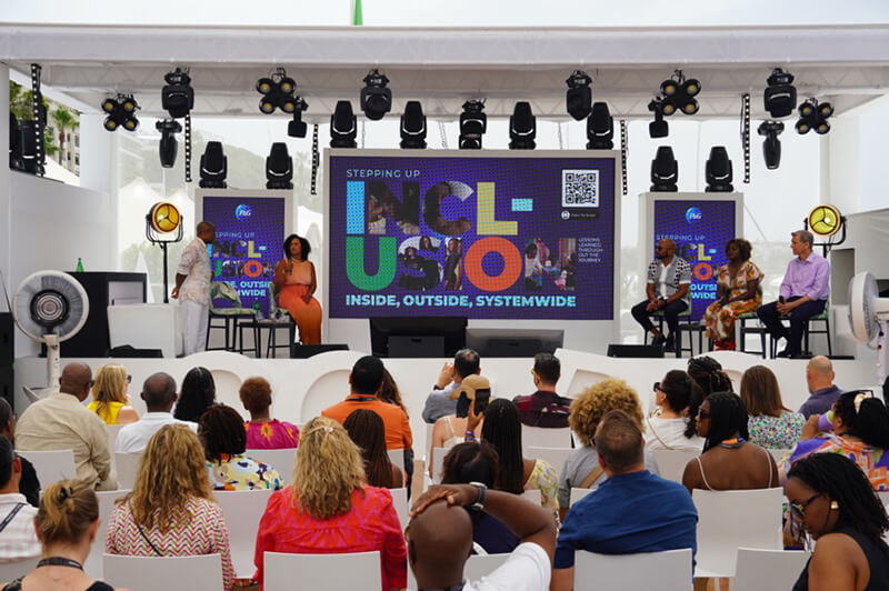 Two black women, two black men and one white man dressed in business attire sit together on a stage. Rows of people in the audience are watching.