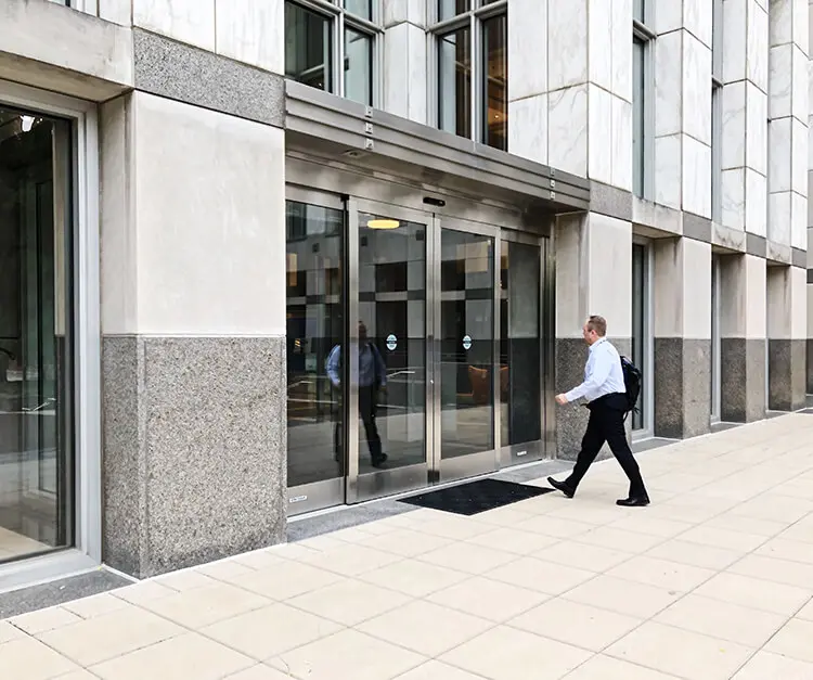 A man walks toward automatic sliding glass doors of a large, concrete office building.