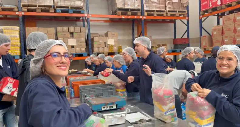 Several men and women wearing hairnets assemble bags of food and relief supplies inside a warehouse. Pallets and boxes are stacked on shelves in the background.