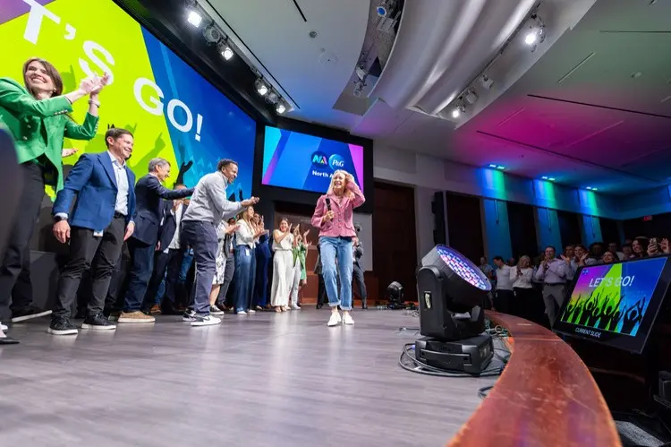 A small crowd of men and women clap and cheer on a blond-haired woman in jeans and a pink jacket as she walks across a stage.