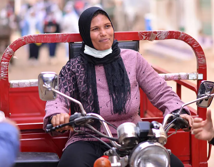 A woman wearing a black head scarf, pink long sleeved shirt and black pants, drives a scooter with a bright red cargo attachment.