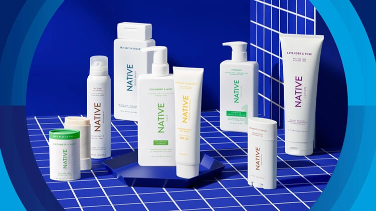 A staggered line-up of Native personal care products sit on top blue square tiles. The white bottles include the brand's logo and product information in various colors.