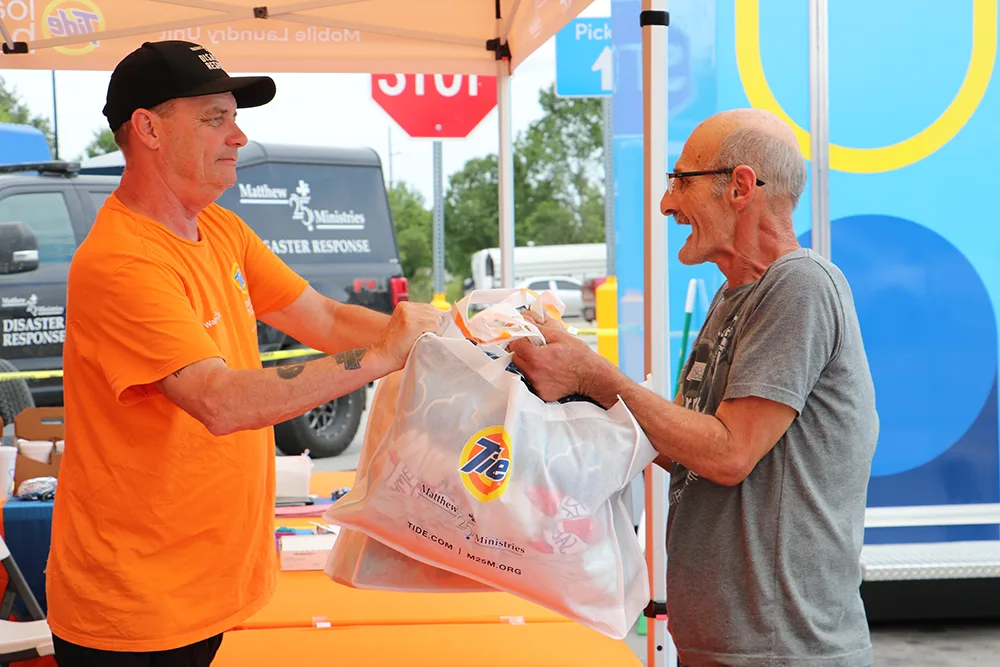 A man in an orange shirt hands a white Tide-branded bag filled with clothes to an older man in a gray shirt, both smiling under an outdoor tent.