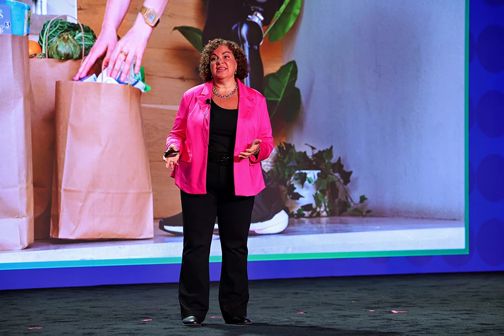 A woman with curly brown hair in a pink blazer standing on stage. She is in front of a screen displaying a person with a prosthetic leg picking up groceries.