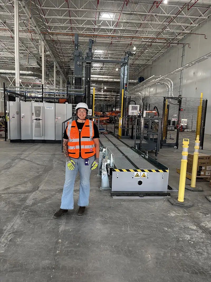 A woman wearing an orange safety vest, hard hat, and gloves stands in a large industrial warehouse with machinery.