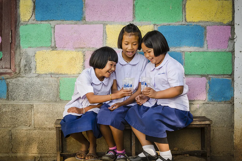 Three young girls with short black hair in blue school uniforms sit on a bench. They laugh as they hold cups of water. Behind them is a colorful brick wall