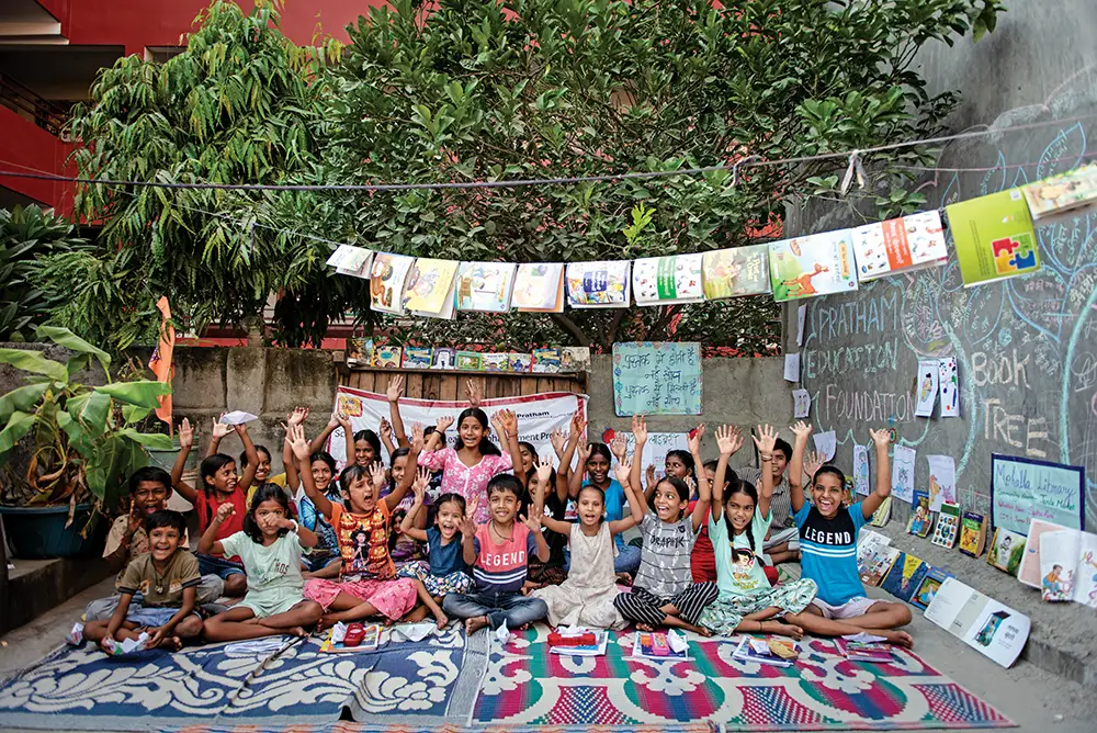 A large group of children sit on colorful mats outdoors with their hands raised, surrounded by hanging books, drawings, and chalkboard art against a garden backdrop.