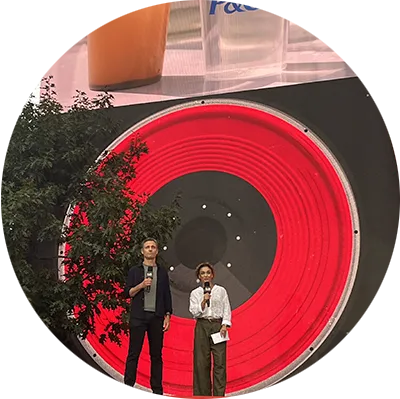A man and woman stand on an outdoor stage. A bright red digital screen is in the background, and a photo of a cup with water.