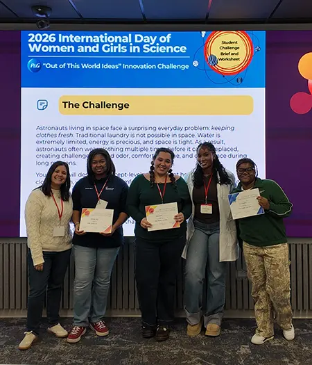 Five young women pose in front of a screen with “The Challenge” written on it in black with a yellow highlight. Some of them hold white certificates in their hands.