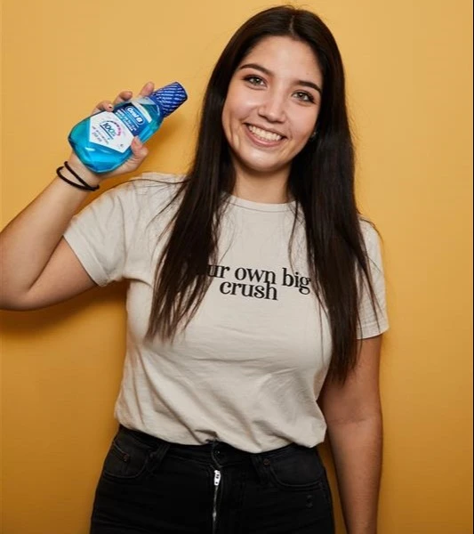 A smiling woman in a light t-shirt and dark jeans holds a blue bottle of Oral-B mouthwash.