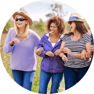 Three women walk arm in arm outdoors on a sunny day, laughing together. They are casually dressed in clothes of various shades of blue.