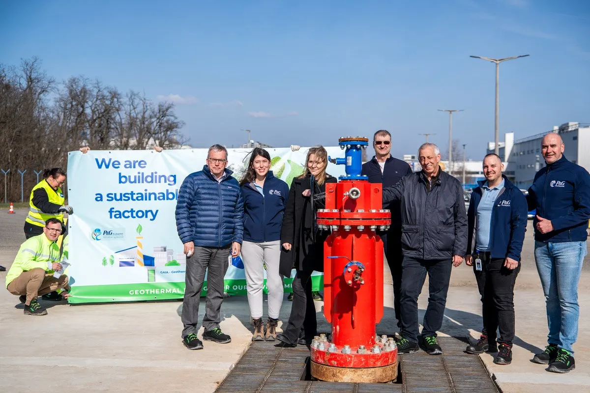 Photograph of several people standing outdoors around a large red industrial valve, with a banner in the background reading "We are building a sustainable factory."